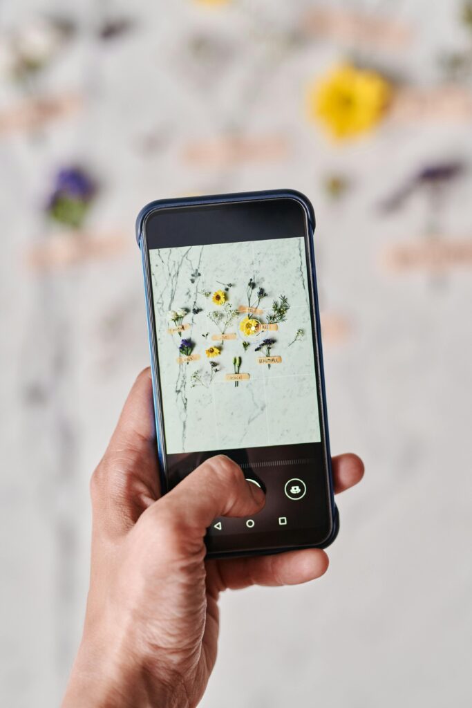 A hand holding a smartphone taking a photo of flowers on a marble surface.